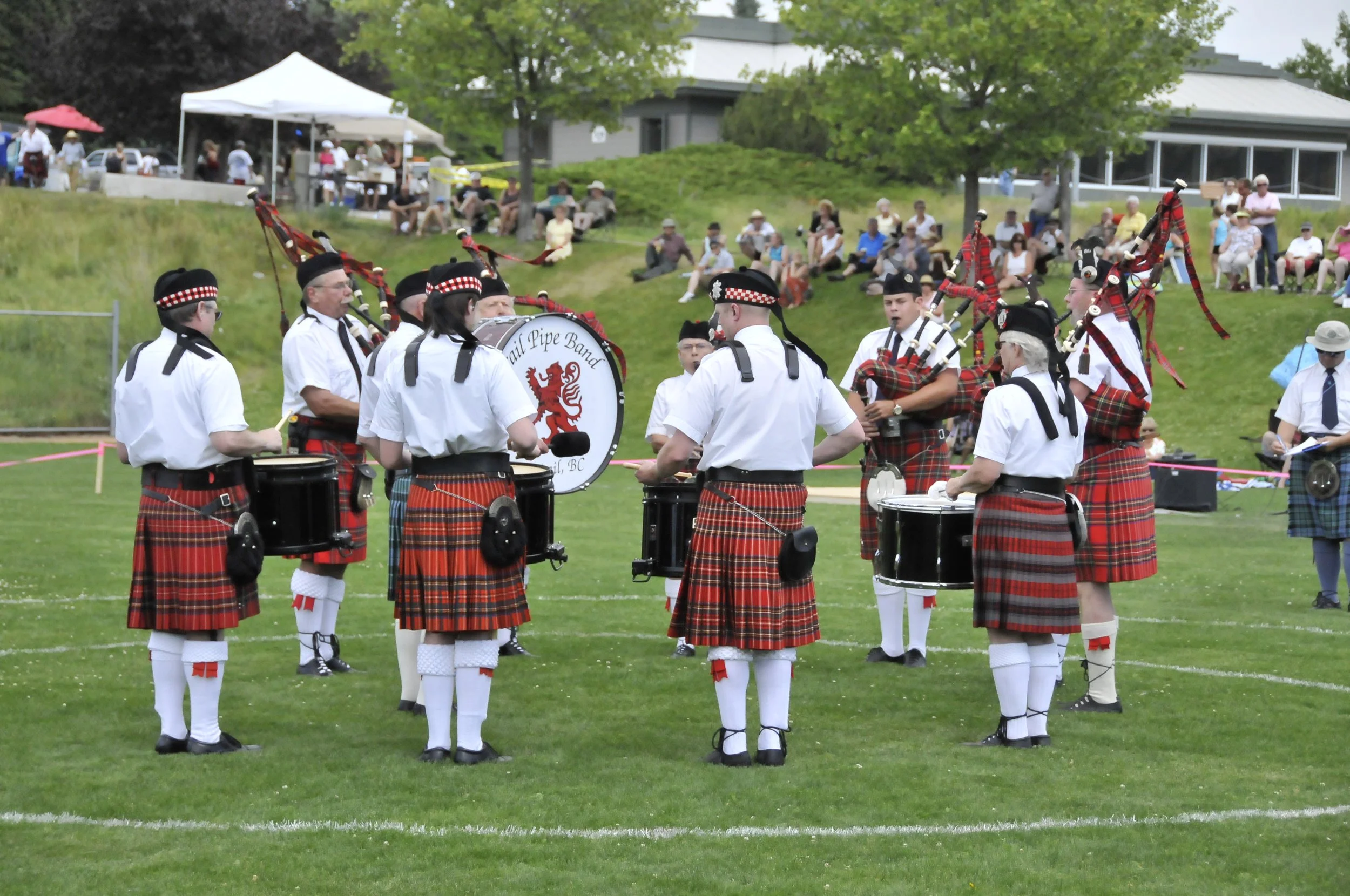 A Scottish pipe band performing outdoors on a grassy field, with members wearing traditional tartan kilts, white shirts, and black hats. Some members are playing drums, while others play bagpipes. An audience is sitting on a hill behind them, watchin