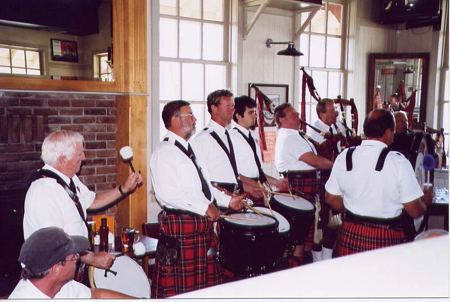 A group of men wearing white shirts, black ties, and tartan kilts, playing traditional Scottish bagpipes and drums inside a cozy pub with brick and wooden decor, large windows, and framed pictures.