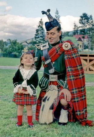 A young girl and a man dressed in traditional Scottish Highland dress, posing outdoors on a grassy field with trees and houses in the background.