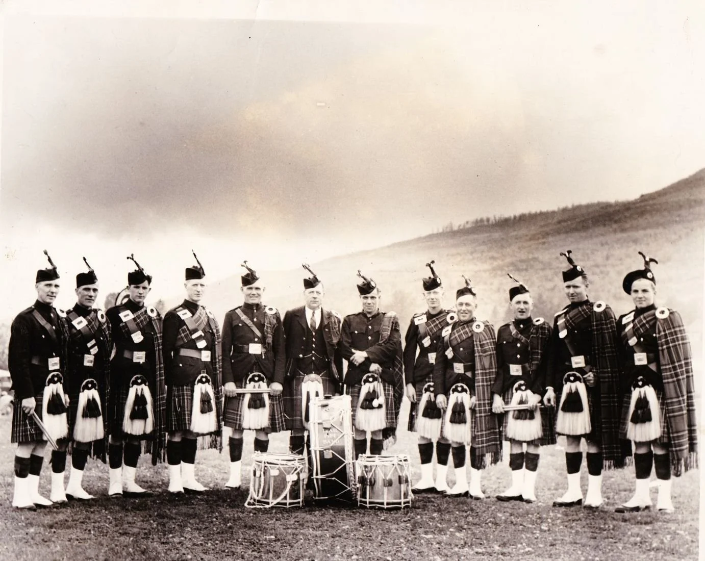 Group of men in traditional Scottish Highland dress with kilts, bagpipes, and drums outdoors, possibly a pipe band, standing on a grassy field with hills in the background.