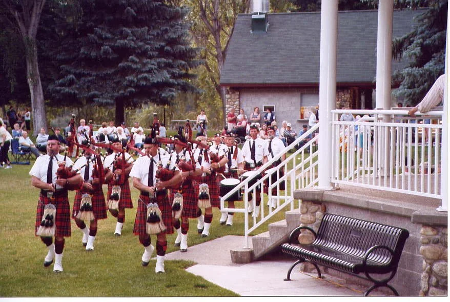 A pipe band dressed in traditional Scottish attire, including kilts, performing outdoors on a grassy area surrounded by trees and a small crowd of spectators.