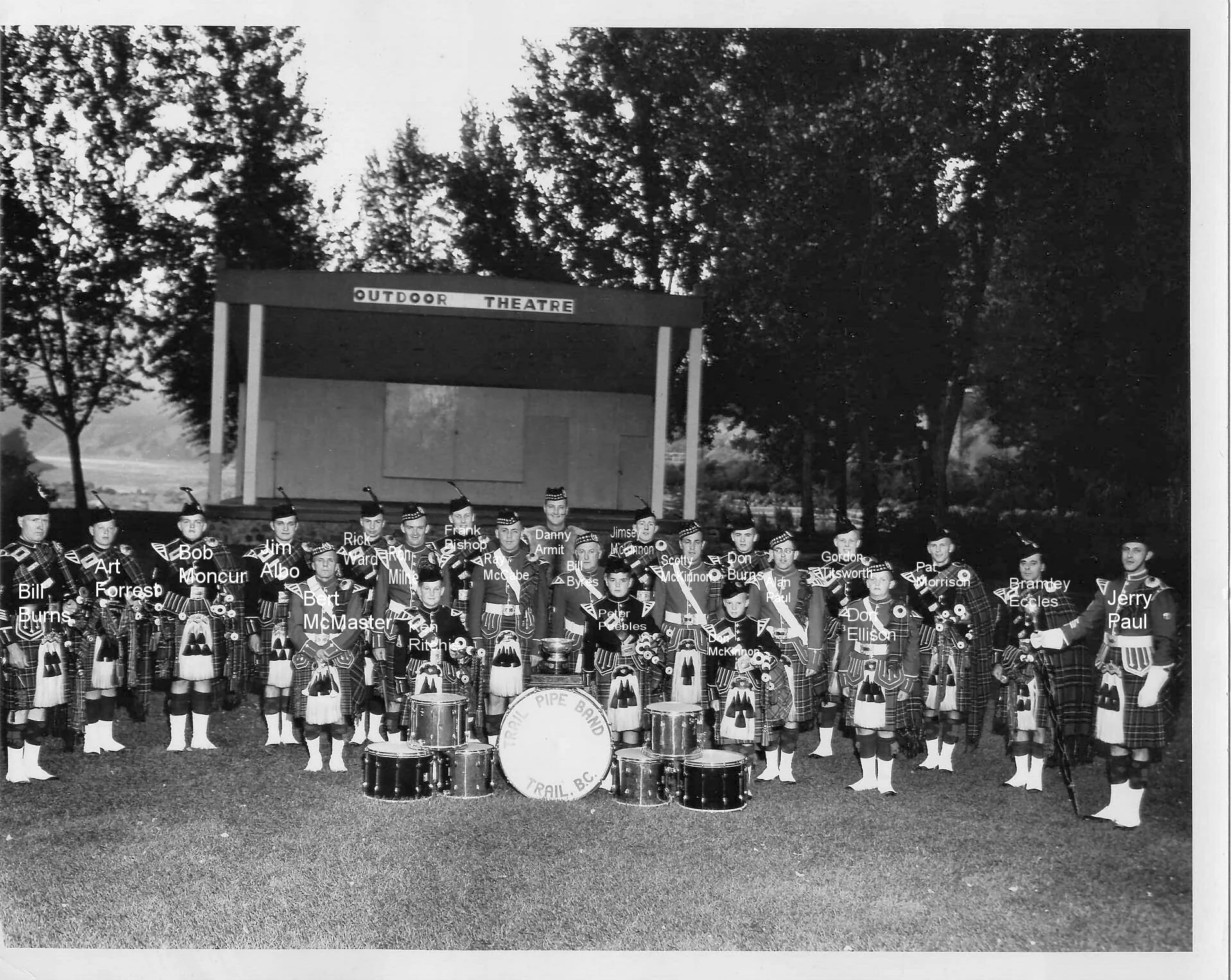 Black and white photo of a pipe band outdoor performance with band members in kilts and hats, standing in front of a stage labeled "Outdoor Theatre" and trees in the background. Names of band members are labeled above them.