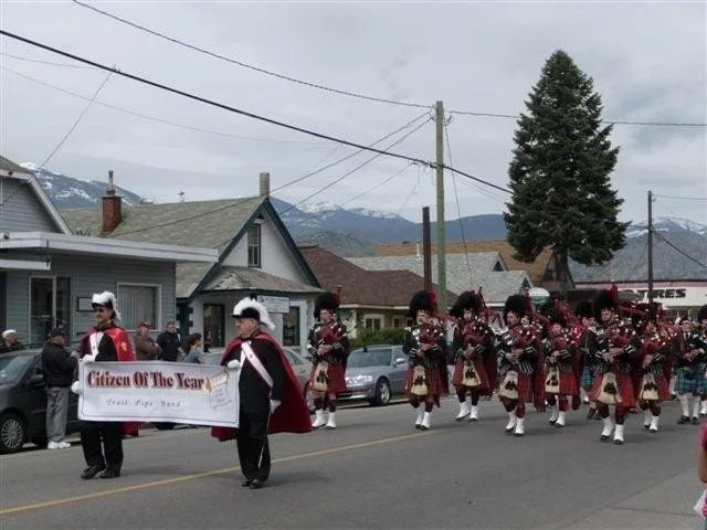 A parade with people dressed in Scottish attire, including kilts and bagpipes, marching down a street with houses and mountains in the background.