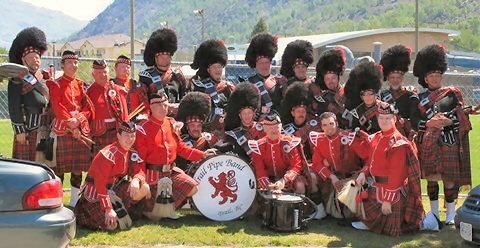A group of marching band members in red uniforms and black bearskin hats posing outdoors with their instruments, and a drum with a lion emblem.