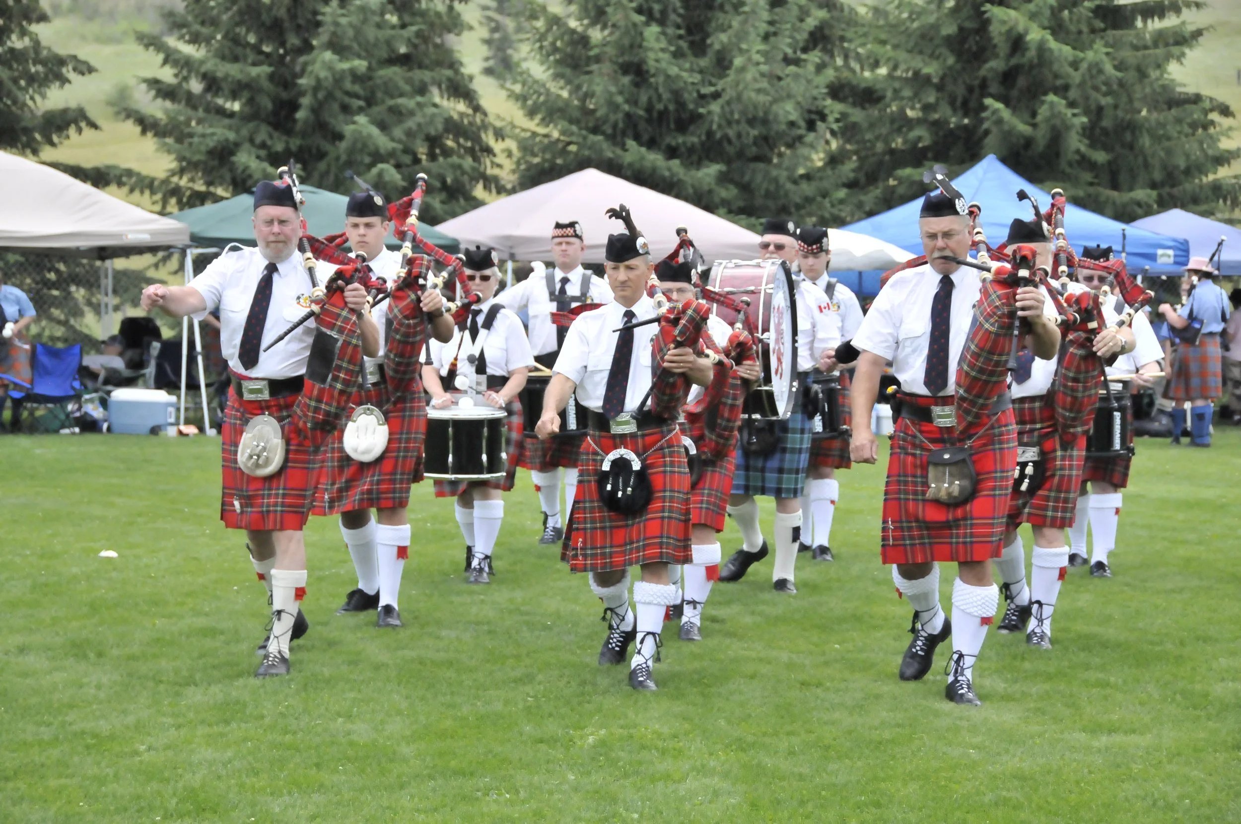 Group of people dressed in traditional Scottish attire, playing bagpipes and drums on a grassy field during a parade or celebration, with tents and spectators in the background.
