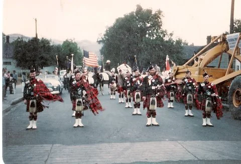 A parade featuring people dressed in traditional Scottish kilts and playing bagpipes, with a construction vehicle in the background.