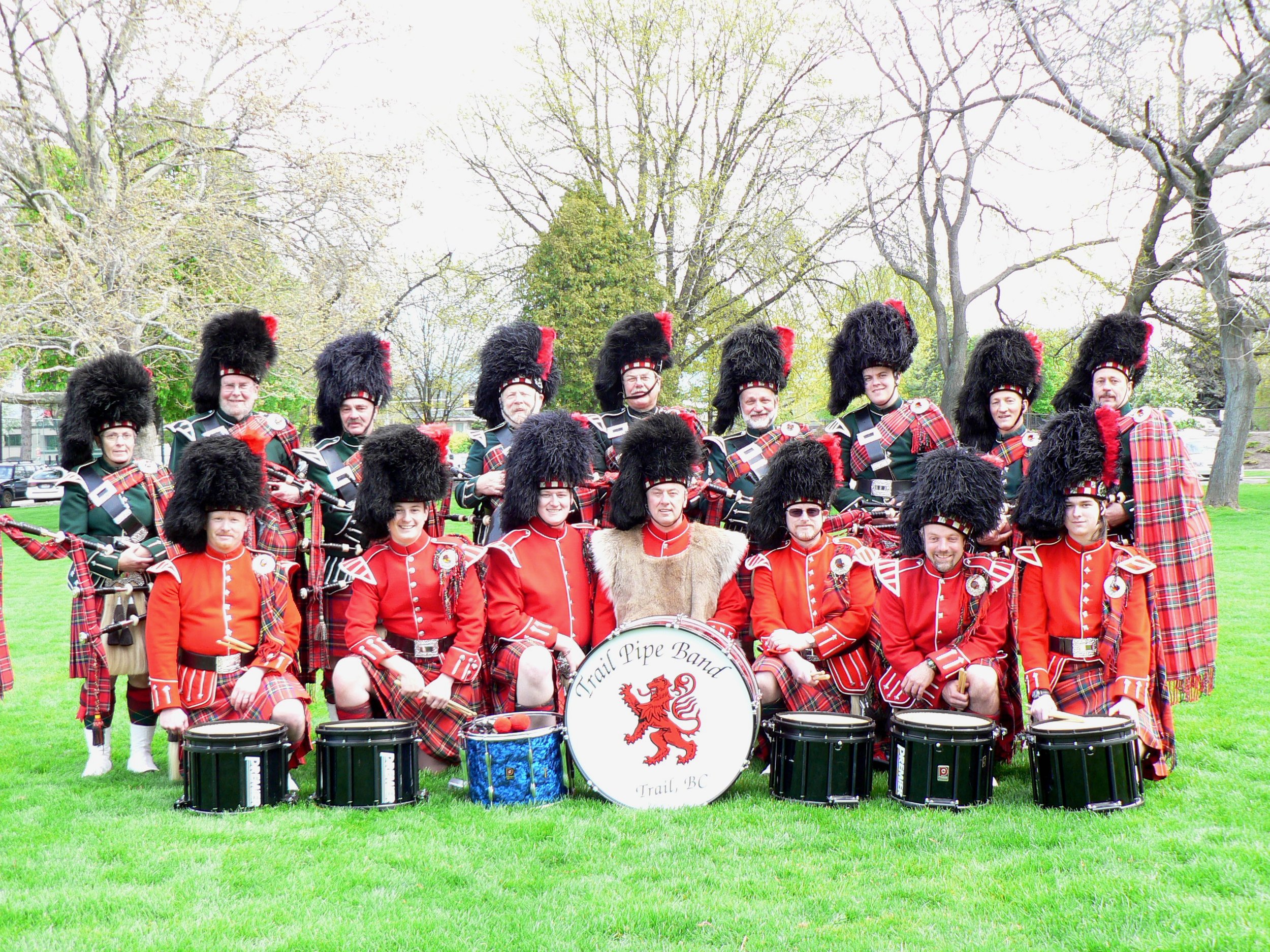 A group of band members dressed in traditional Scottish attire, including kilts and tall black fur hats, posing outdoors on a grassy field with trees in the background. The band's drum features a red lion and the text "Trail Pipe Band Trail, BC.".