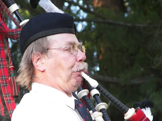 A man playing the bagpipes outdoors, wearing glasses, a black hat, and a white shirt with a necktie, with trees in the background.