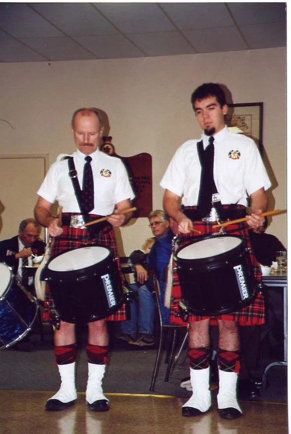 Two men dressed in traditional Scottish attire playing drums at an indoor event with seated attendees in the background.