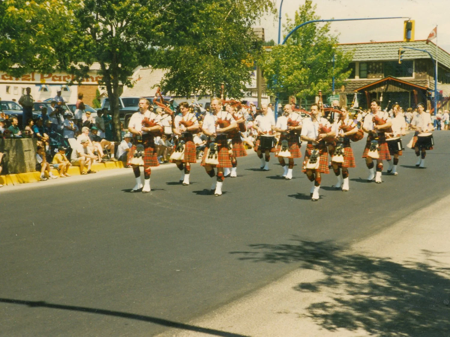 A parade with bagpipers and drummers dressed in traditional Scottish kilts, marching on a street lined with spectators.