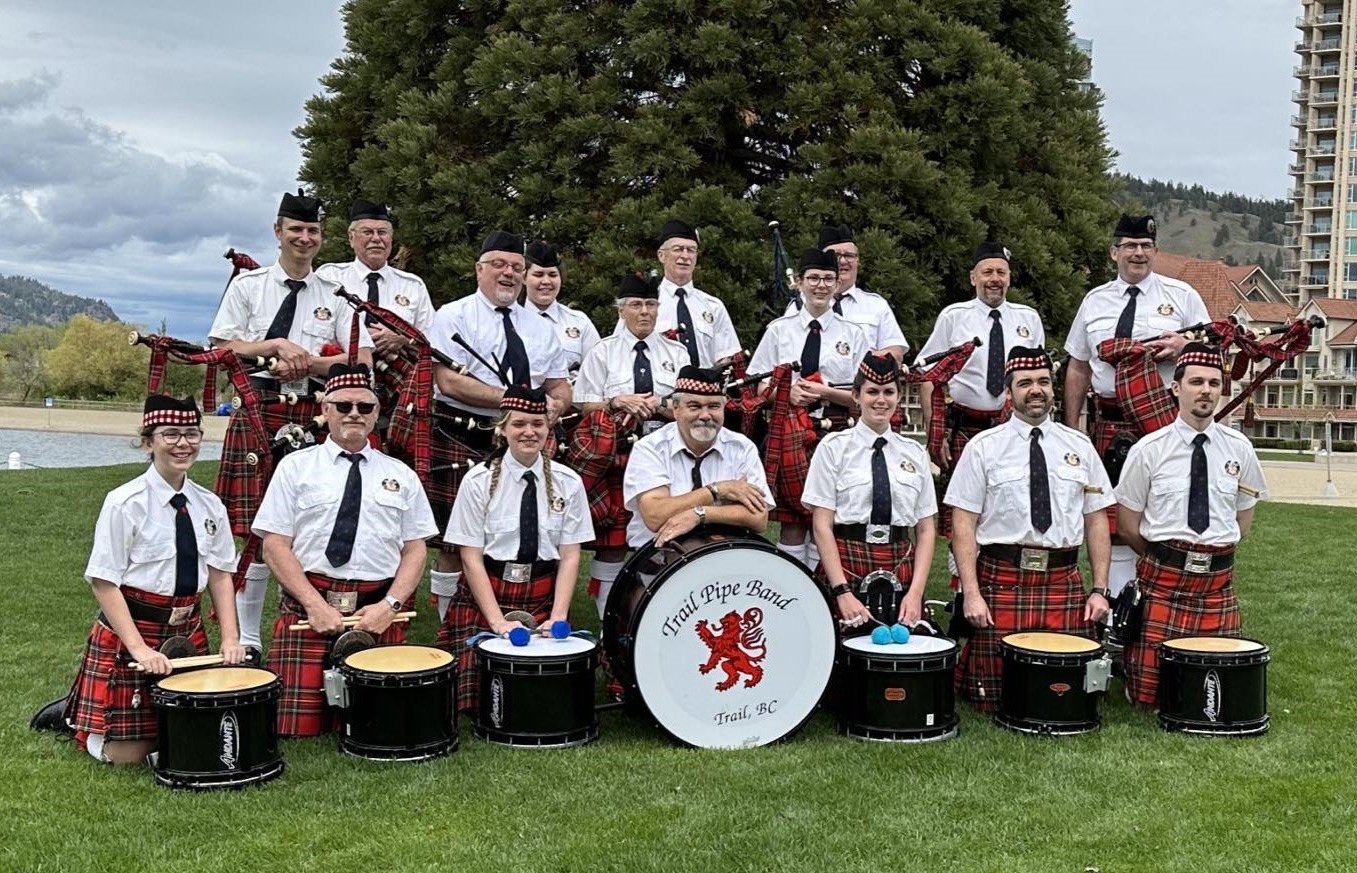 A group of 14 people in matching uniforms, some holding bagpipes, posing outdoors with a large tree and buildings in the background. They are members of the Trail Pipe Band, Trail, BC, standing on grass.
