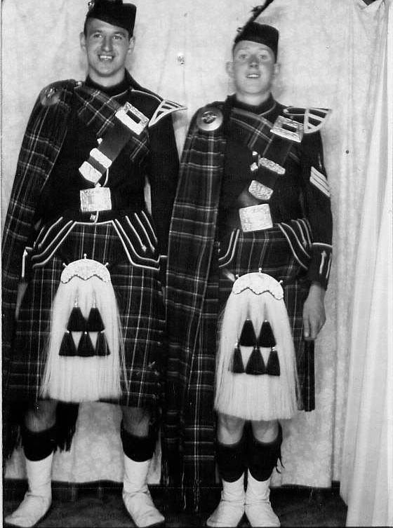 Two men dressed in traditional Scottish Highland dress, including kilts, sporrans, and jackets, standing side by side. They are smiling and standing in front of a plain curtain backdrop.