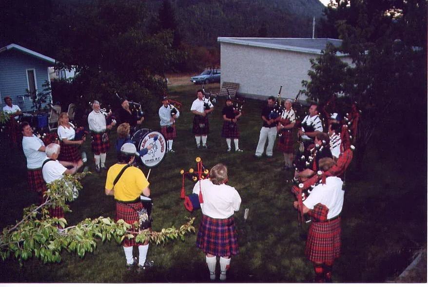 A group of people in traditional Scottish attire, playing bagpipes and drums in a circle outdoors, with trees, a house, and a car in the background.