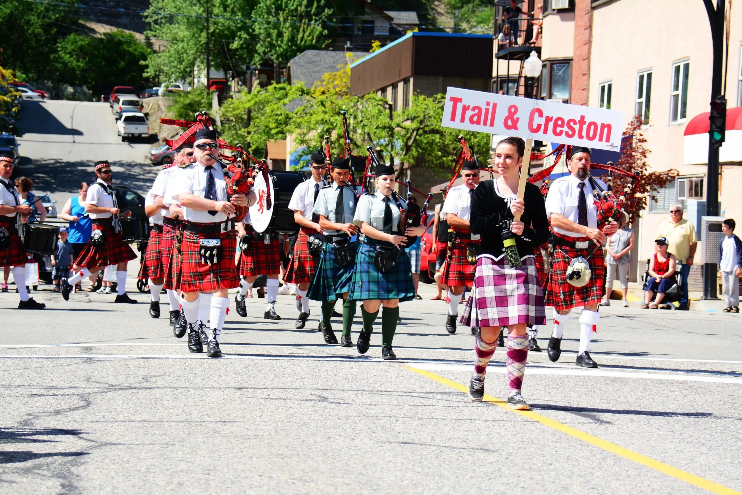 A parade with people in traditional Scottish attire. The group is marching on a street, some holding musical instruments and a woman at front holding a sign that reads "Trail & Creston." Spectators are watching from the side of the street, with trees