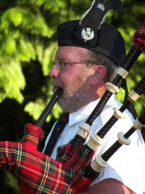 A man in traditional Scottish attire playing the bagpipes outdoors, wearing a black hat with a white feather and a white shirt, with green trees in the background.