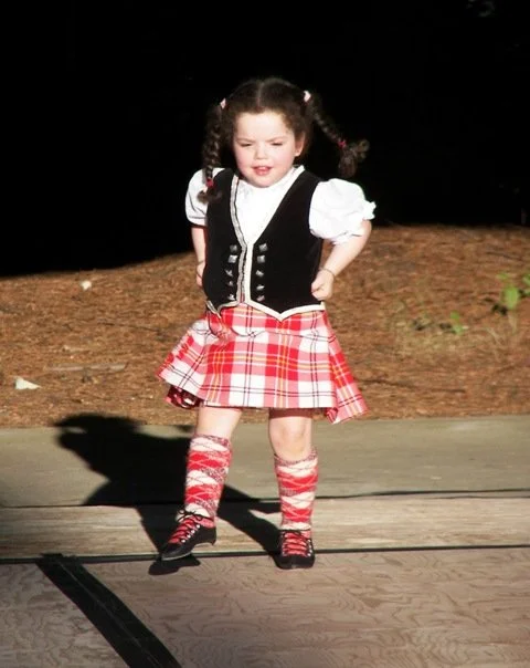 A young girl with dark hair in pigtails, wearing a black vest, white blouse, red plaid skirt, and matching knee-high socks, standing outdoors on a wooden surface with her hands on her hips.