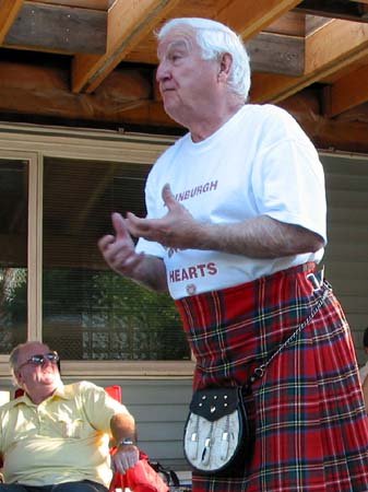 An elderly man wearing a white t-shirt and a tartan kilt standing and speaking, with a man in sunglasses sitting nearby and listening, under a wooden porch.