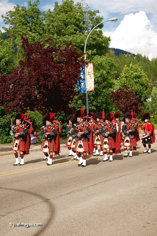 A group of bagpipe players dressed in traditional Scottish attire, including kilts and tall hats, performing during a parade on a street lined with green trees and a cloudy sky.