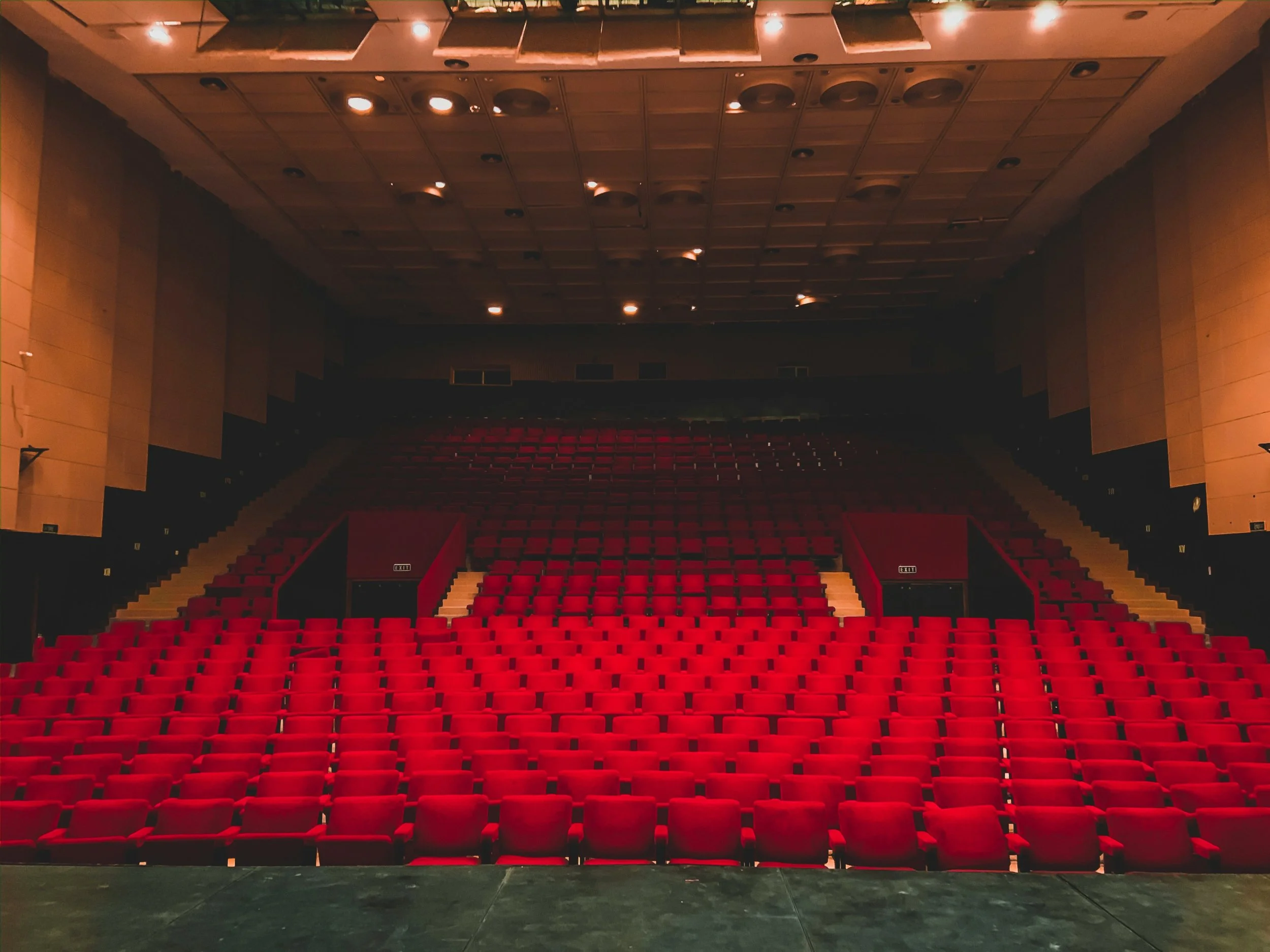 A theater auditorium with rows of empty red seats viewed from the stage. The warm lighting highlights the seating and architectural details. The space feels grand and ready for an audience.