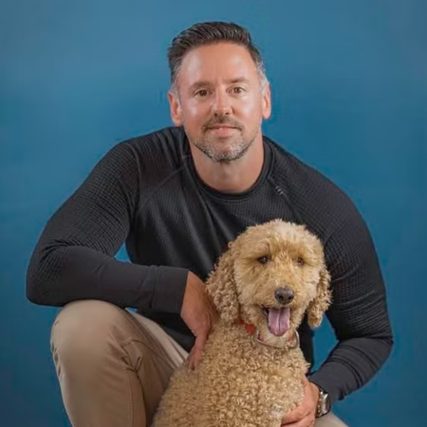 A man with short gray hair and a beard, wearing a black long-sleeve shirt and beige pants, kneels next to a curly-haired tan dog with a collar, both posing in front of a blue background.