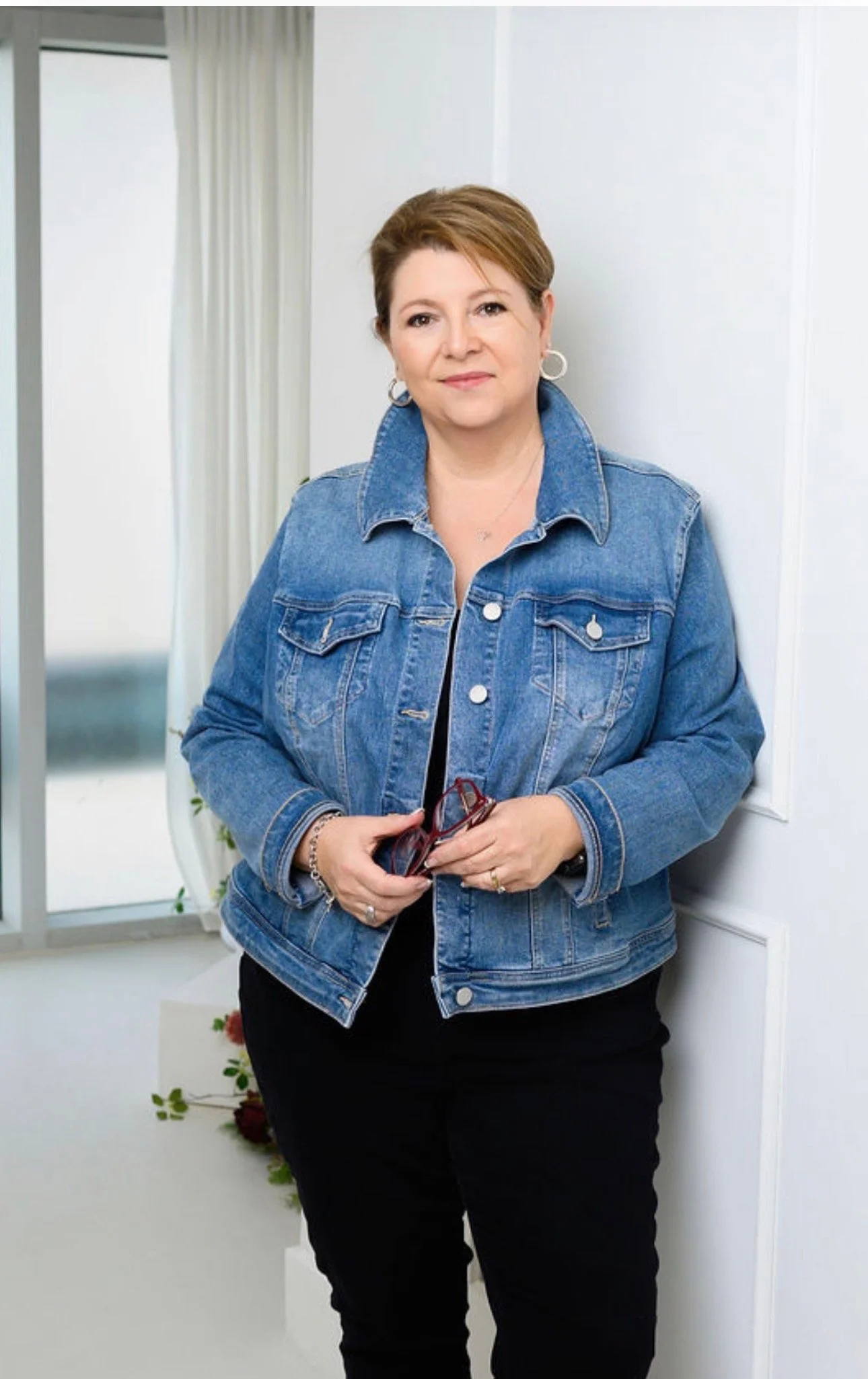 Professional woman in black blazer and dress sitting on the arm of a black couch, smiling, with a gray wall background and the text 'Aileen Araujo Sales Representative' in the top left corner.