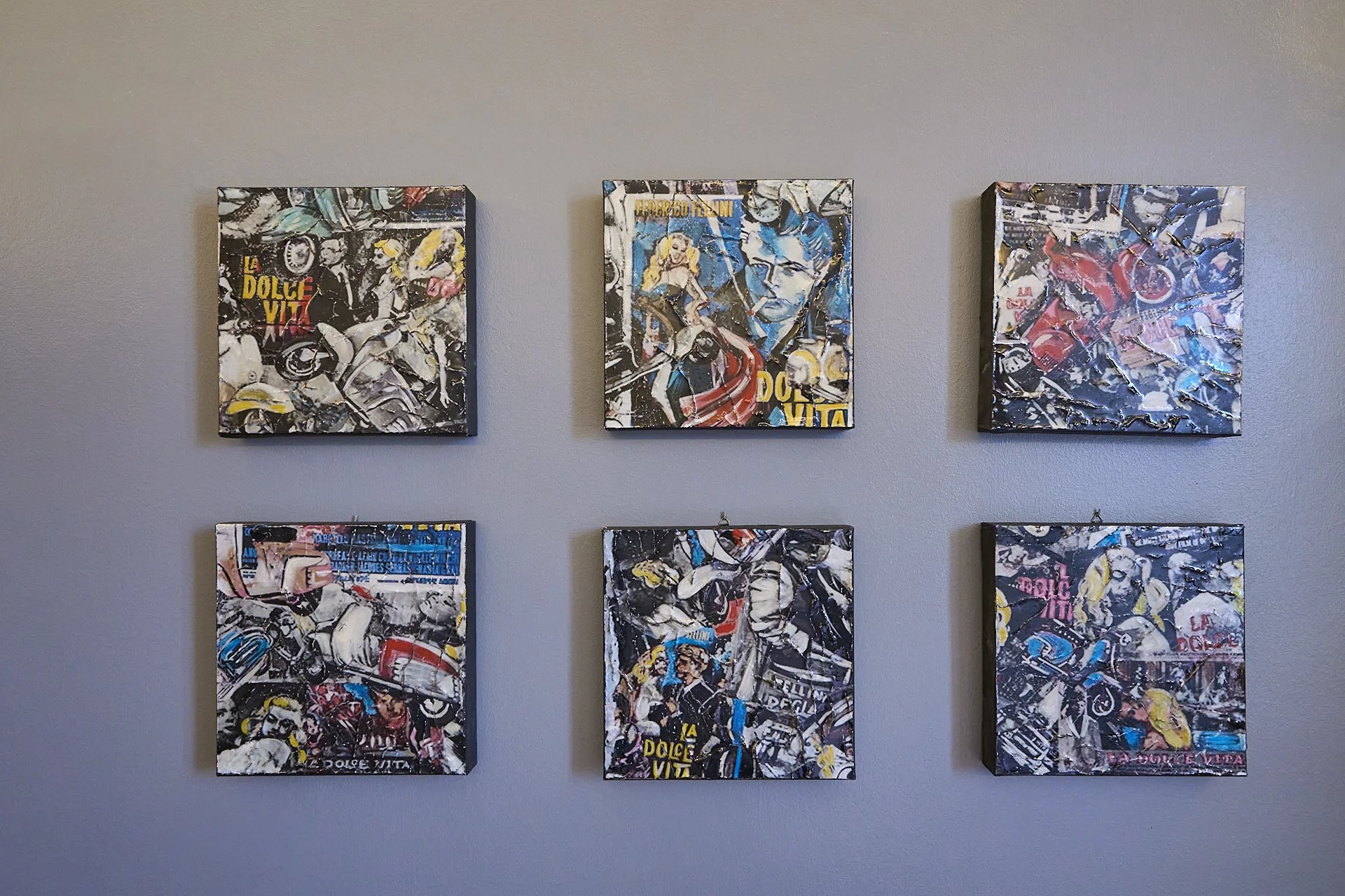 Sei quadri astratti con immagini di motociclette, persone e testi in italiano, appesi a una parete grigia.