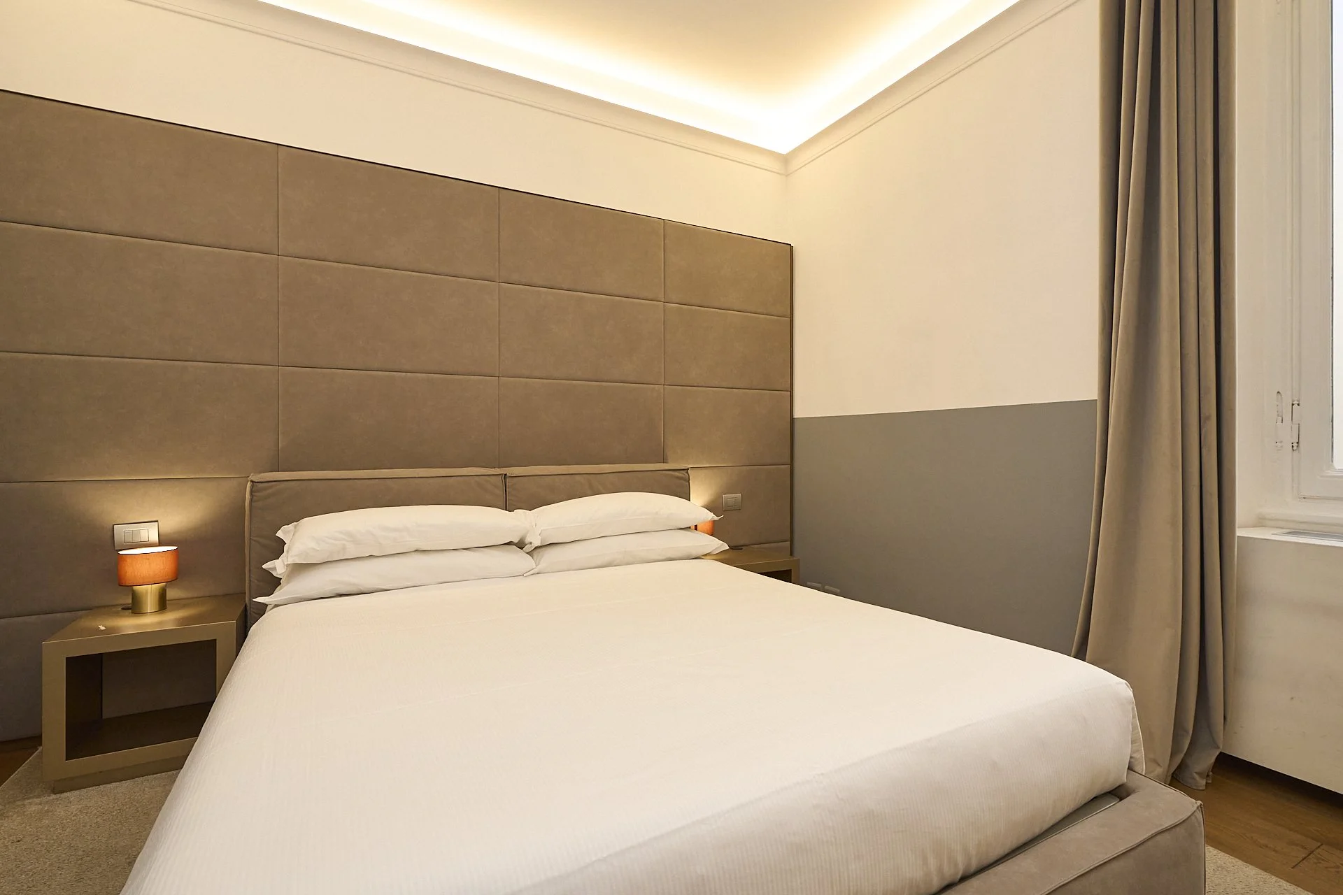 Camera moderna con letto matrimoniale, due comodini con lampade accese, tende beige e pareti chiare, illuminata da luci soffuse.