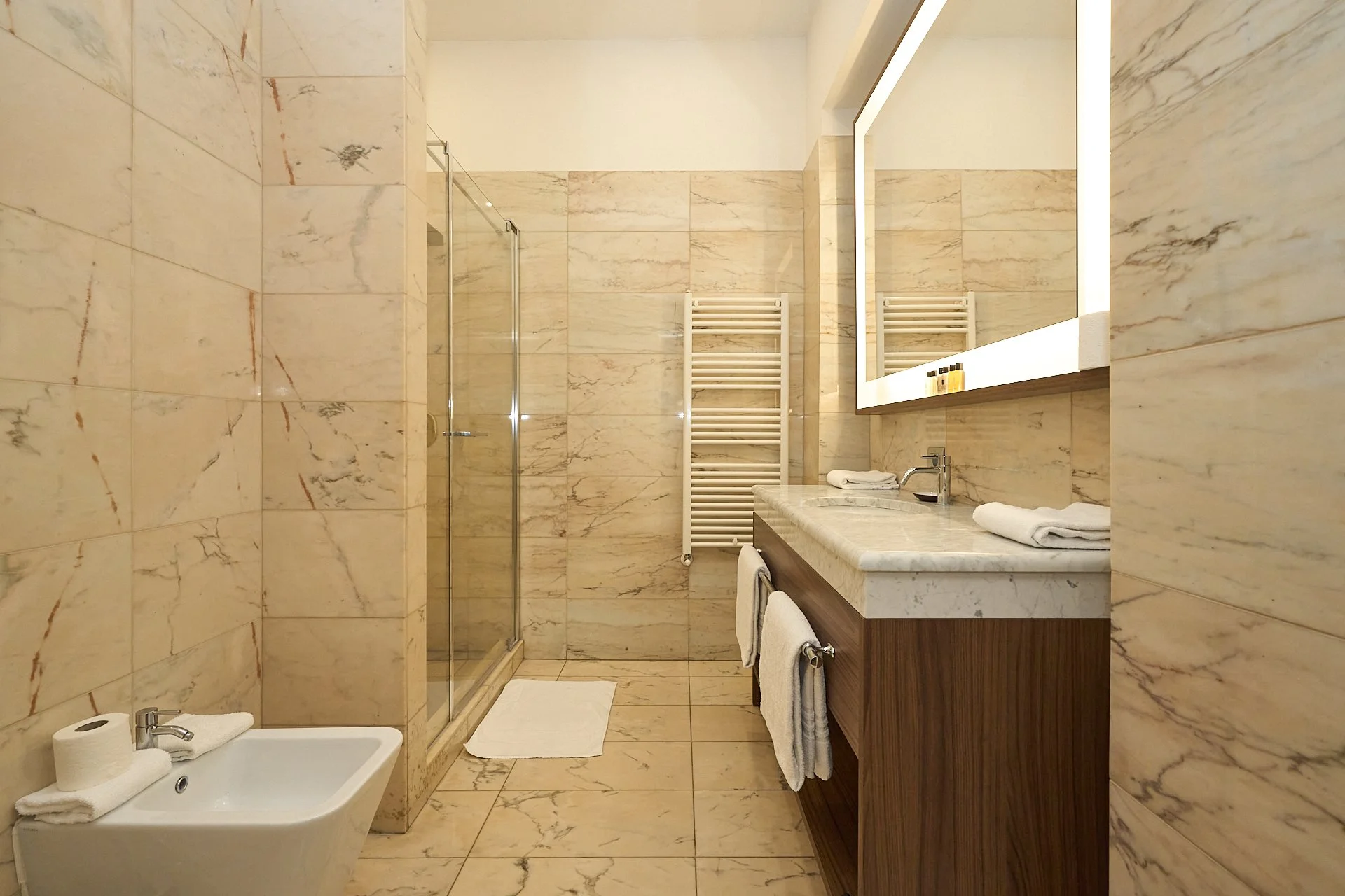 Bagno con pareti e pavimento in marmo beige, lavabo con mobile in legno scuro, grande specchio con luce, doccia con porta in vetro, radiator bianco, lavabo bidet con asciugamani bianchi, tappeto bianco davanti alla doccia.