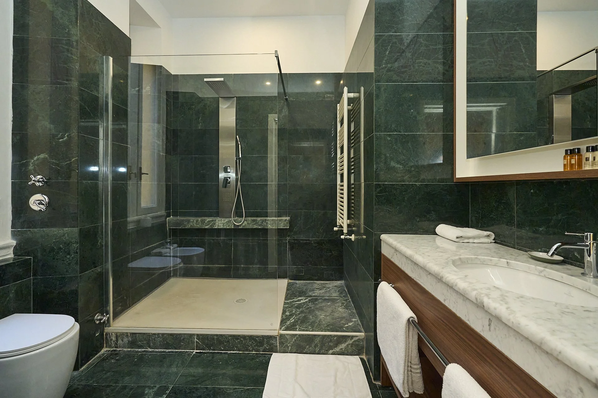 Bagno con pareti e pavimenti in marmo verde, doccia con soffione a soffitto e pannello di controllo, lavandino con grande specchio e piccoli flaconi, asciugamani bianchi.
