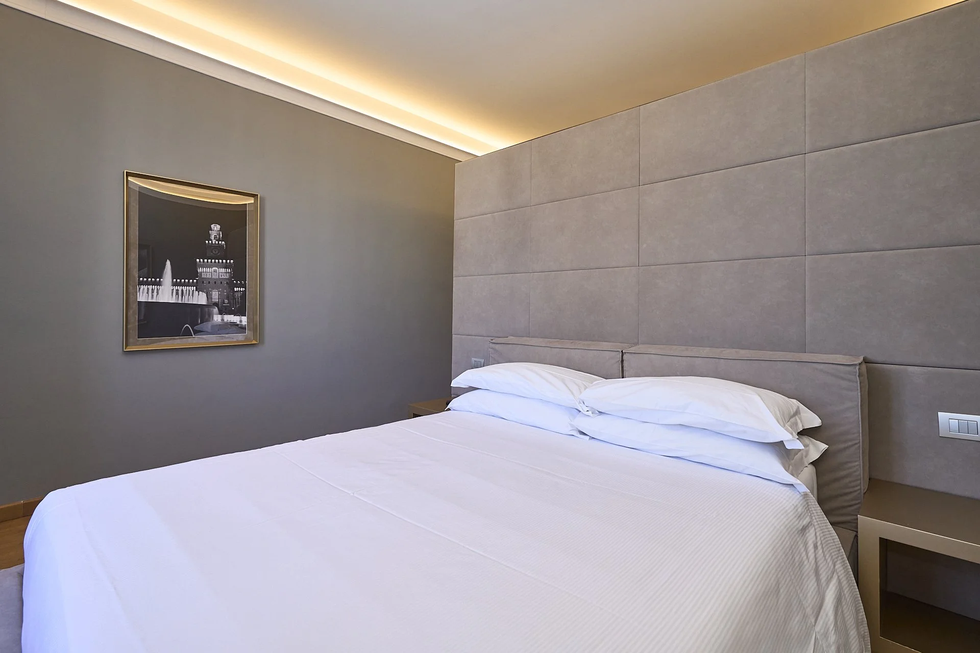 Letto matrimoniale con cuscini bianchi, testata in tessuto beige, pareti grigie con quadro decorativo e angolo della stanza con luce soffusa.