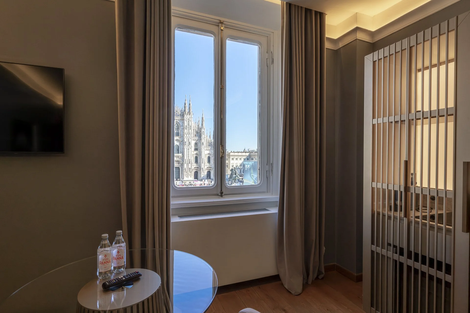 Interno di una stanza con finestra che guarda verso una cattedrale, con tende chiuse e una piccola teca di vetro, due bottiglie d'acqua e un telecomando sul tavolo