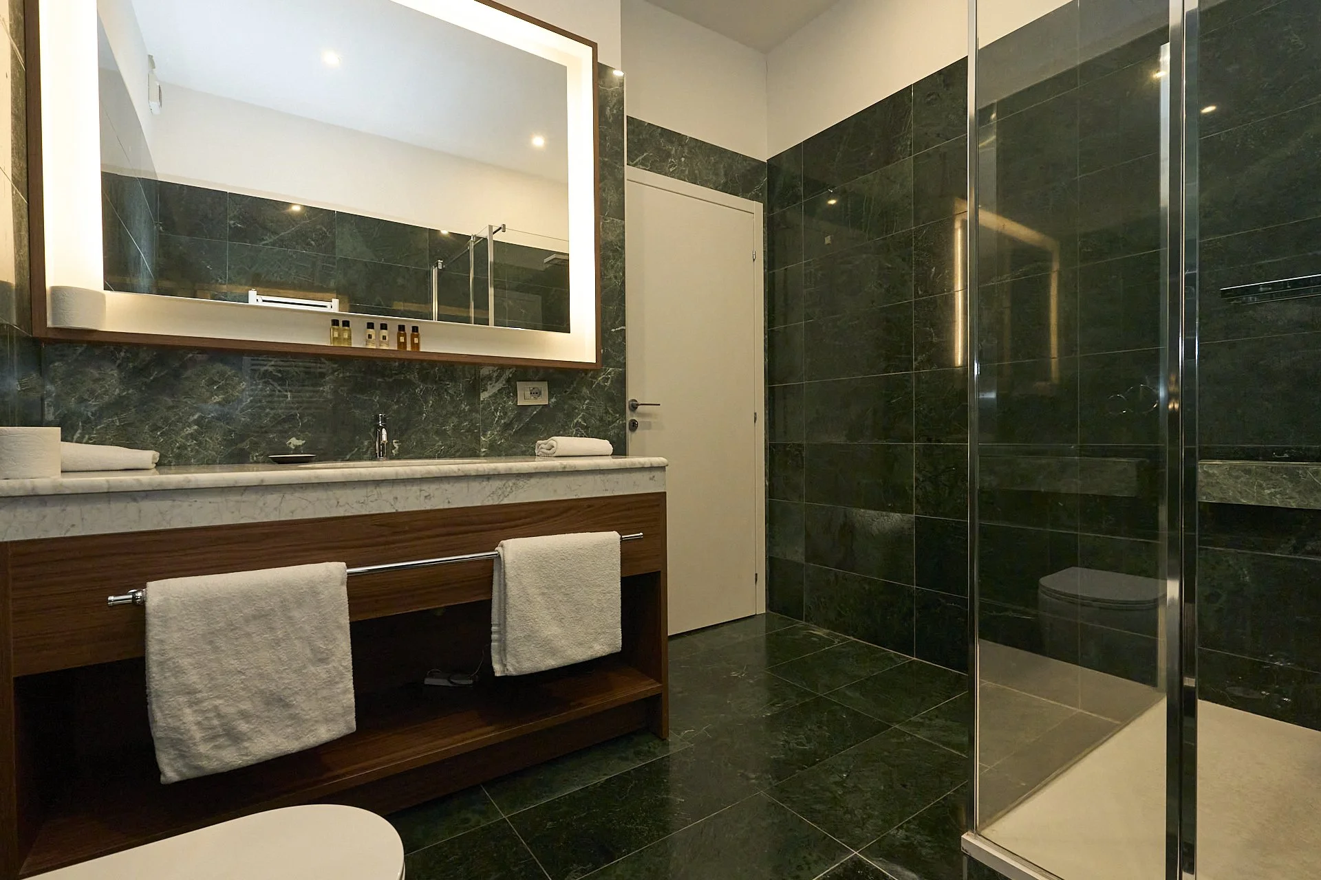 Bagno con pareti e pavimento in marmo verde scuro, grande specchio sopra lavandino, lavandino con rivestimento in marmo, asciugamani bianchi, porta bianca, cabina doccia con porte di vetro, elettrovalvola visibile vicino al lavabo.