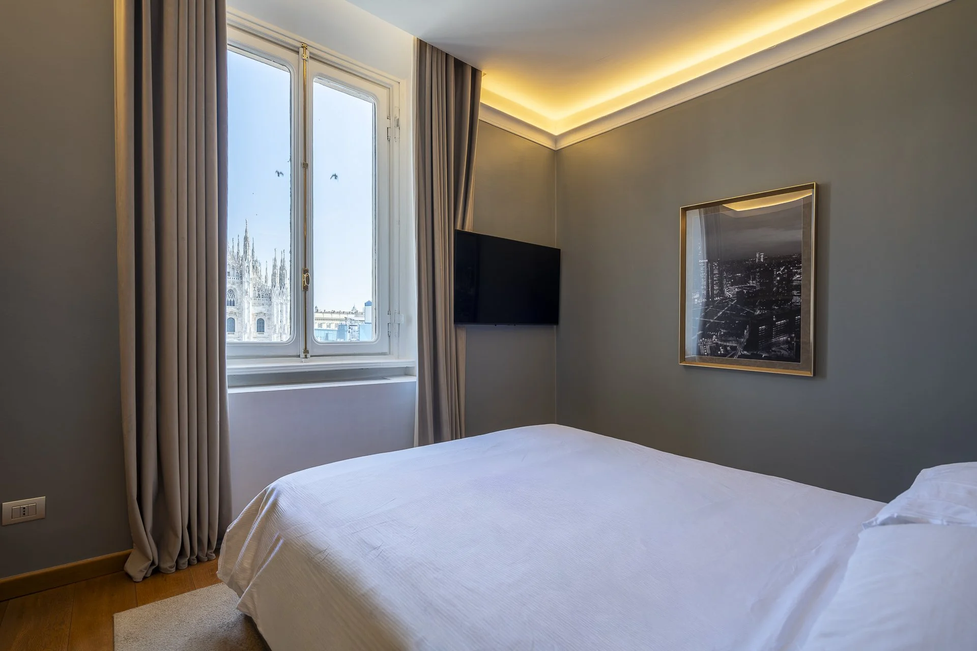 Camera da letto con pareti verdi, finestra con tende beige e vista sulla città, letto con sottomisura bianca, televisore a schermo piatto e quadro con vista urbana di notte.