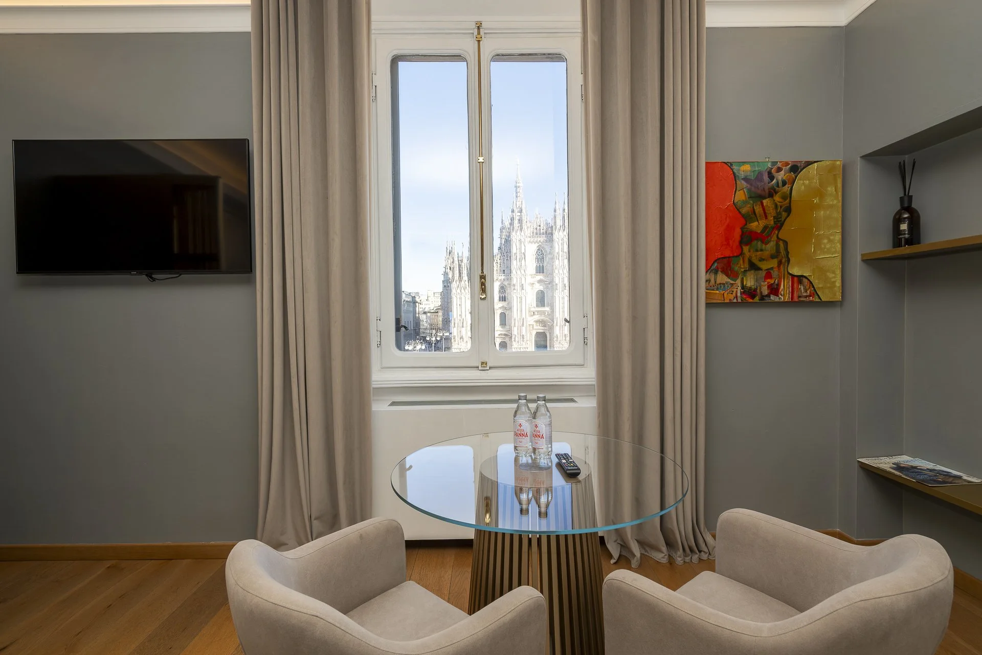 Vista di una stanza con due poltrone beige intorno a un tavolino di vetro, due bottiglie d'acqua e un telecomando. Sullo sfondo, una finestra con vista sulla cattedrale di Milano, con tende chiare e un quadro colorato sulla parete destra.
