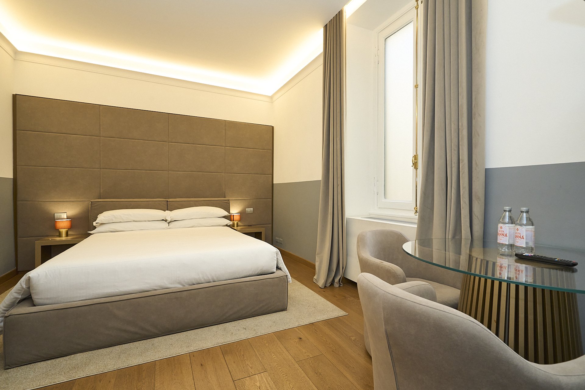 Camera da letto moderna con letto matrimoniale, tappeto beige, due lampade ai lati del letto, grande finestra con tende e sedie di fronte a un tavolo con bottiglie d'acqua e telecomando.