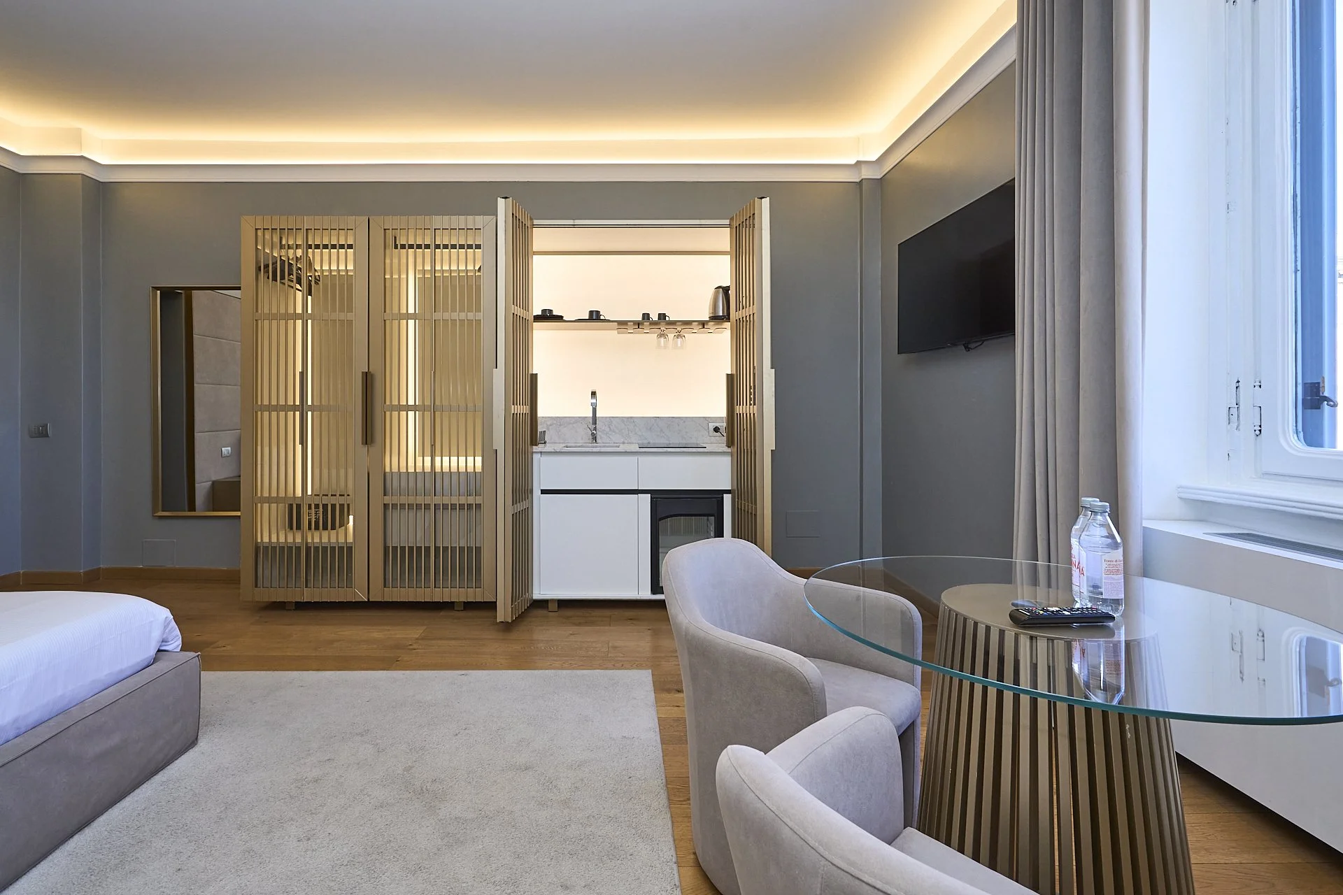 Camera moderna con area soggiorno e angolo cottura, sedia e tavolo di vetro, vista su finestra con tende bianche, televisore a parete, e produzione di luce soffusa dal soffitto.