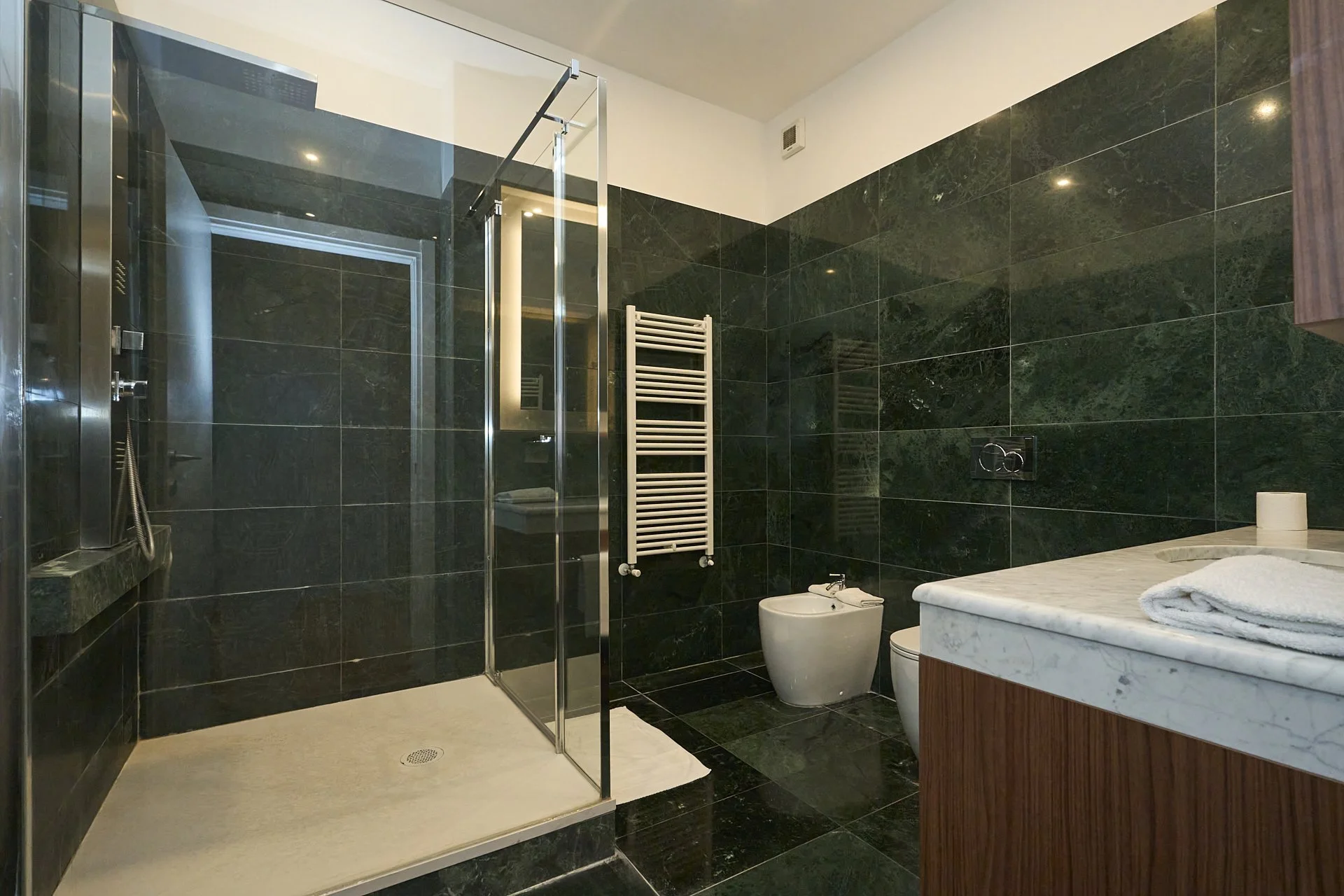 Bagno moderno con pareti e pavimento in ceramica verde scuro, doccia con pareti di vetro, sanitari bianchi e lavabo in marmo con mobile in legno.