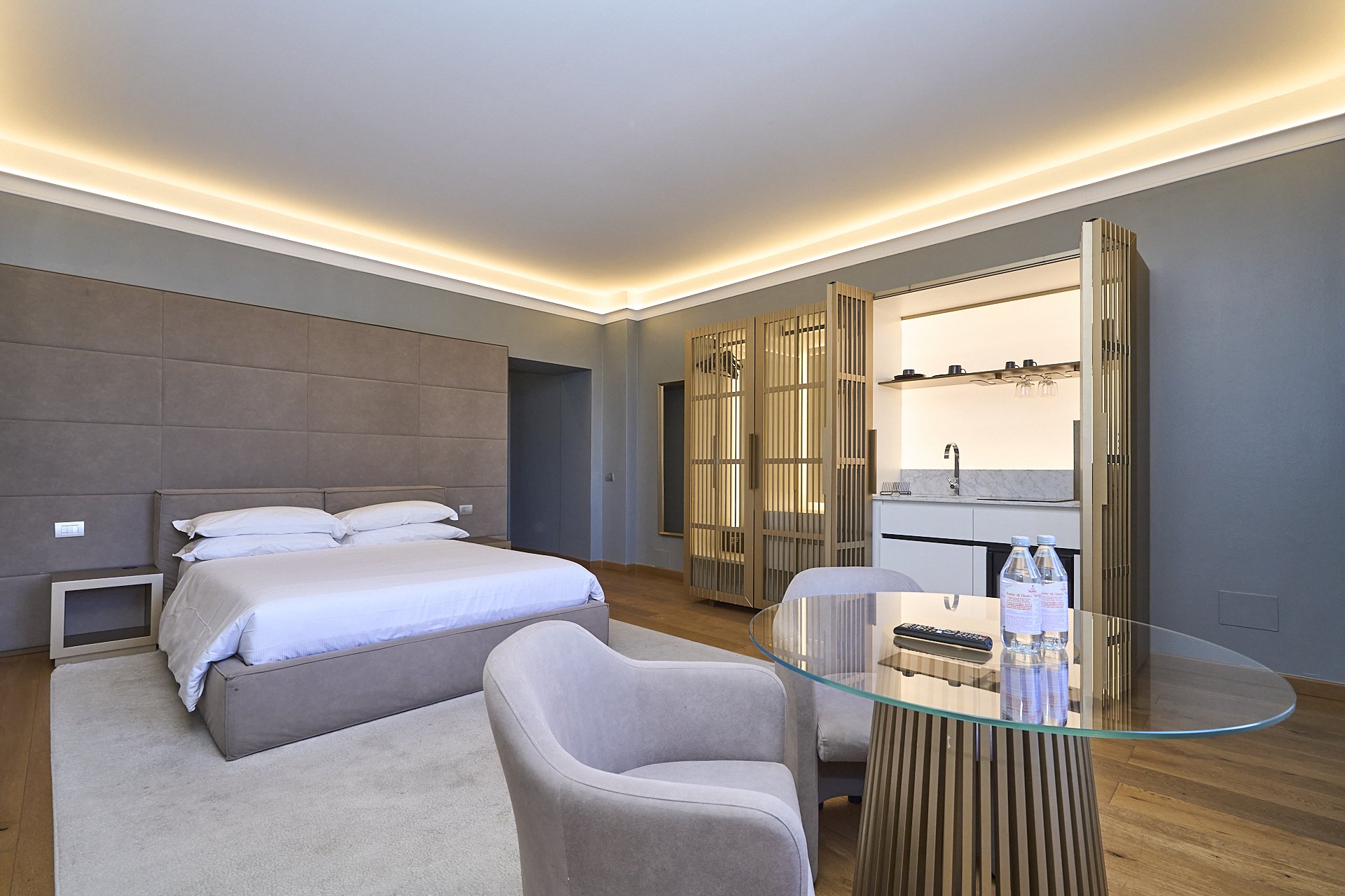 Camera da letto moderna con letto matrimoniale, soggiorno con tavolo di vetro e sedie, piccola cucina con lavabo, acqua e telecomando.