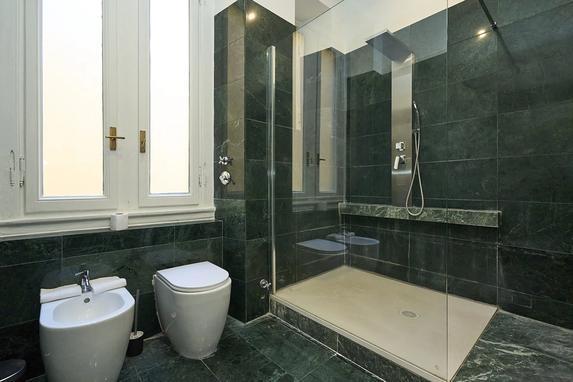 Bagno con pareti e pavimenti in marmo verde scuro, finestra con inferiate, doccia walk-in con parete in vetro e piatto doccia beige, bidet e wc bianco.