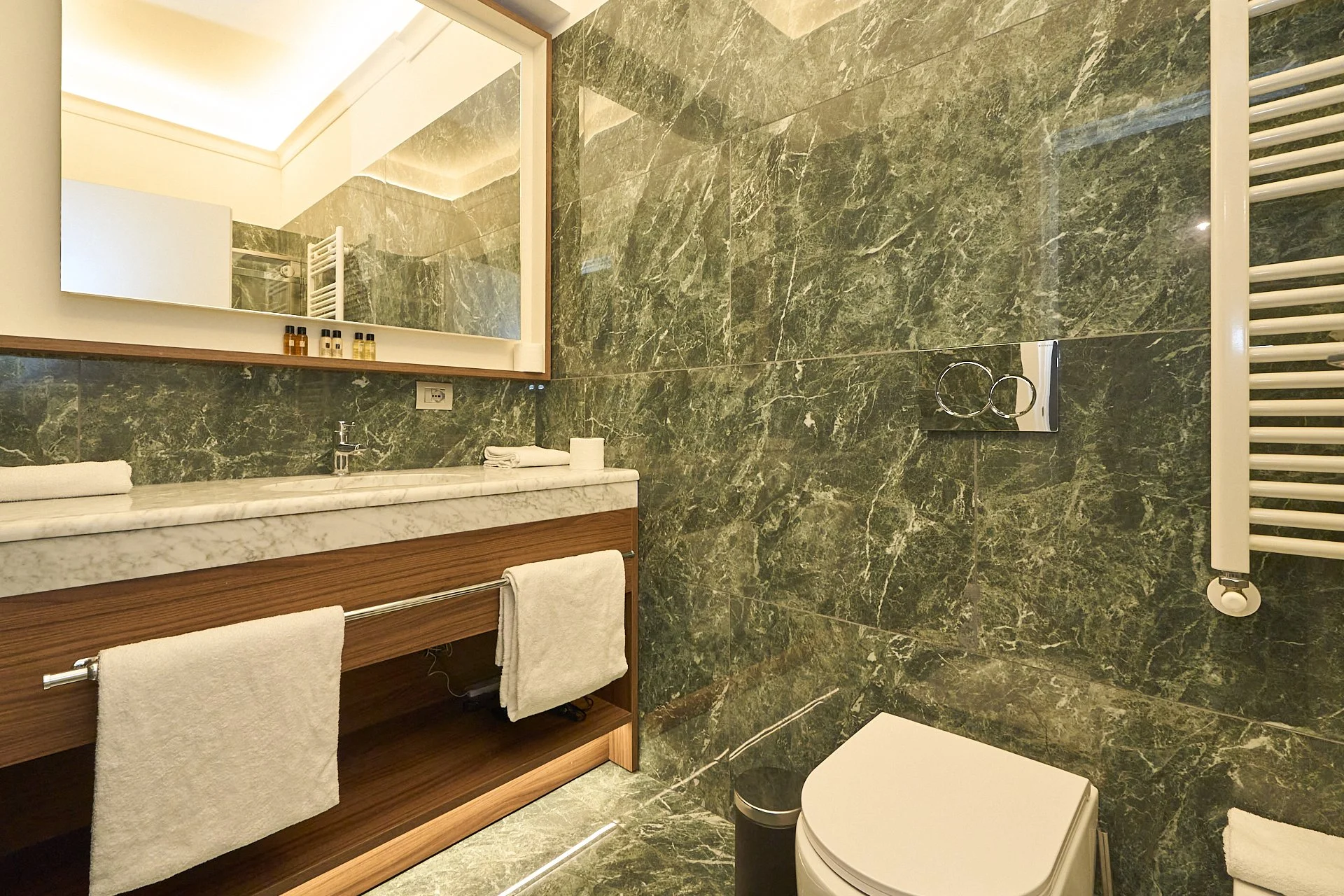 Bagno con pareti e pavimenti in marmo verde scuro, grande specchio sopra il lavabo in marmo bianco, asciugamani bianchi pendono da una barra sotto il lavabo, vaso sanitario bianco, termosifone verticale bianco, piccolo bidoncino nero accanto al water