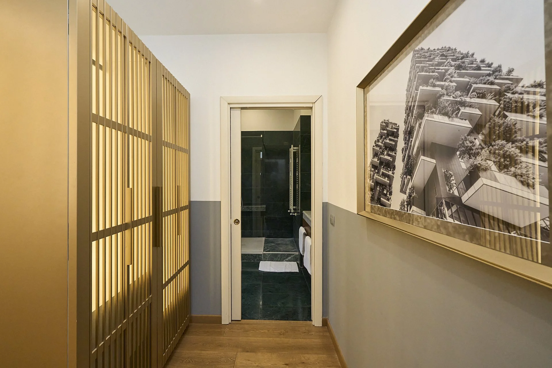 Corridor con armadio a muro in legno, pareti bianche e grigie, e una porta che conduce a un bagno con piastrelle nere e un asciugamano.