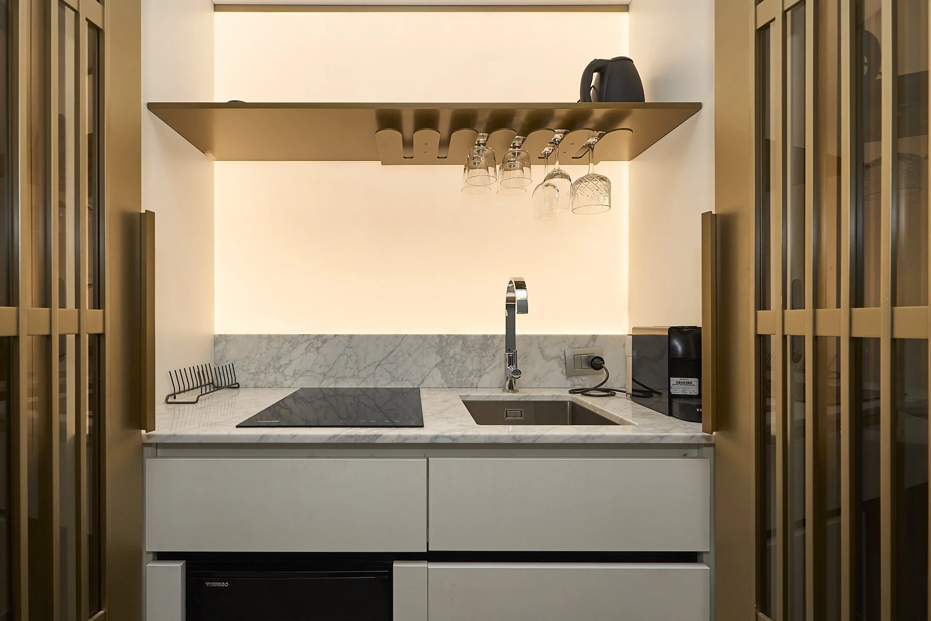 Mini cucina moderna con piano in marmo, lavello e piano di cottura, sopra un mobile con un ripiano di vetro con bicchieri appesi e vari elettrodomestici come moka e microonde.