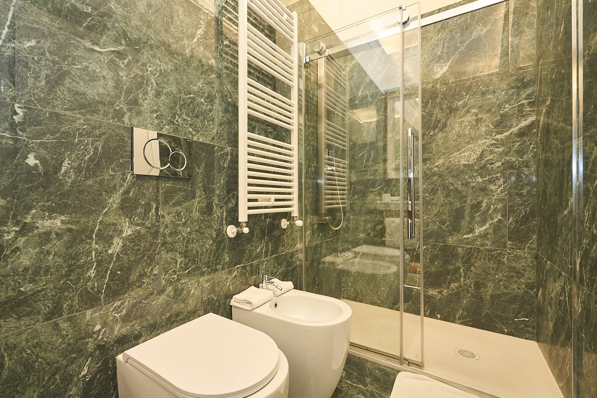 Bagno moderno con pareti e pavimento in marmo verde scuro, doccia con porta in vetro, radiatore bianco e servizi igienici bianchi