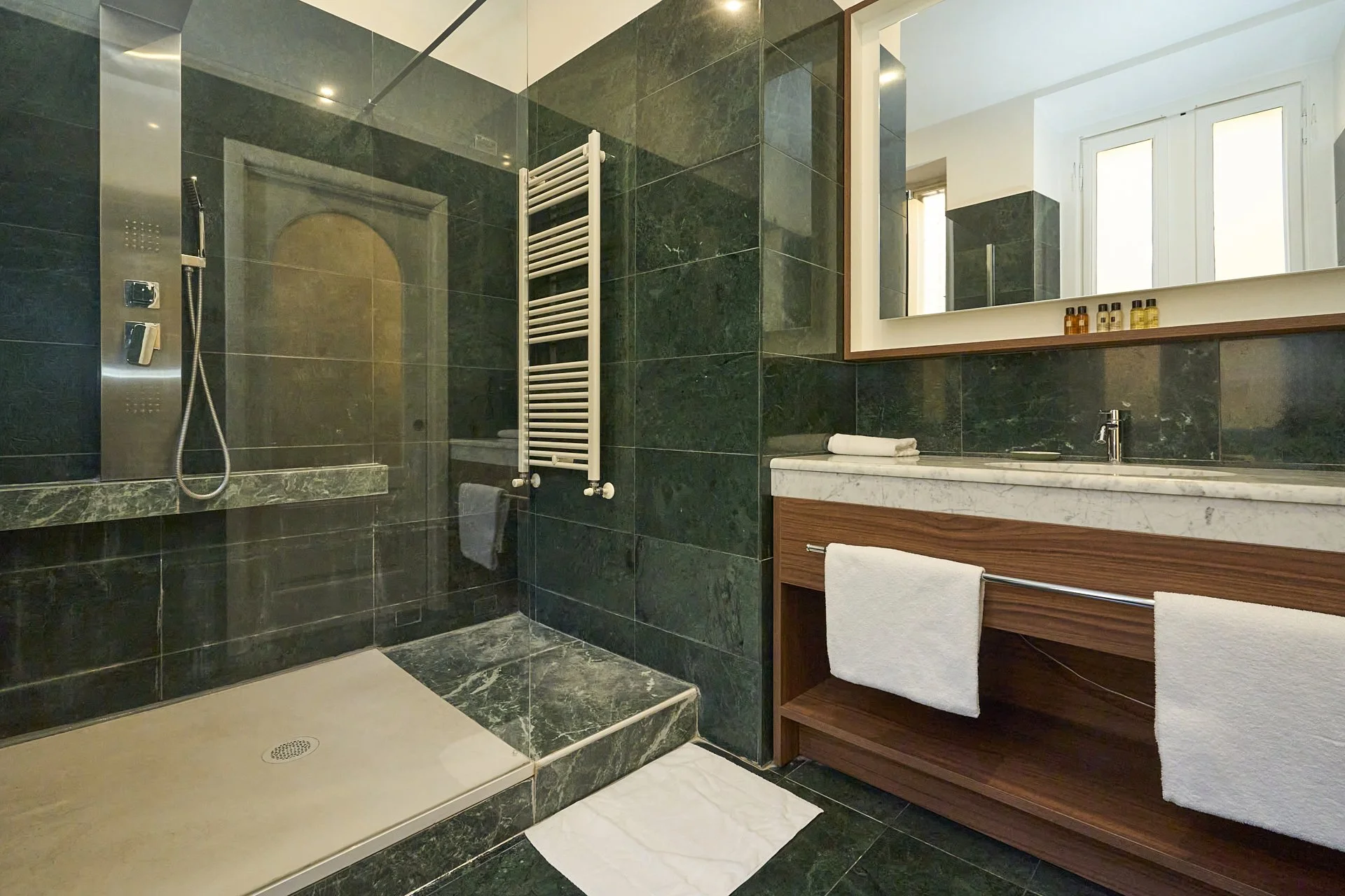 Bagno moderno con pareti e pavimento in piastrelle verdi, doccia con piatto in pietra naturale, grande specchio sopra lavabo in marmo con mobile in legno, e asciugamani bianchi.