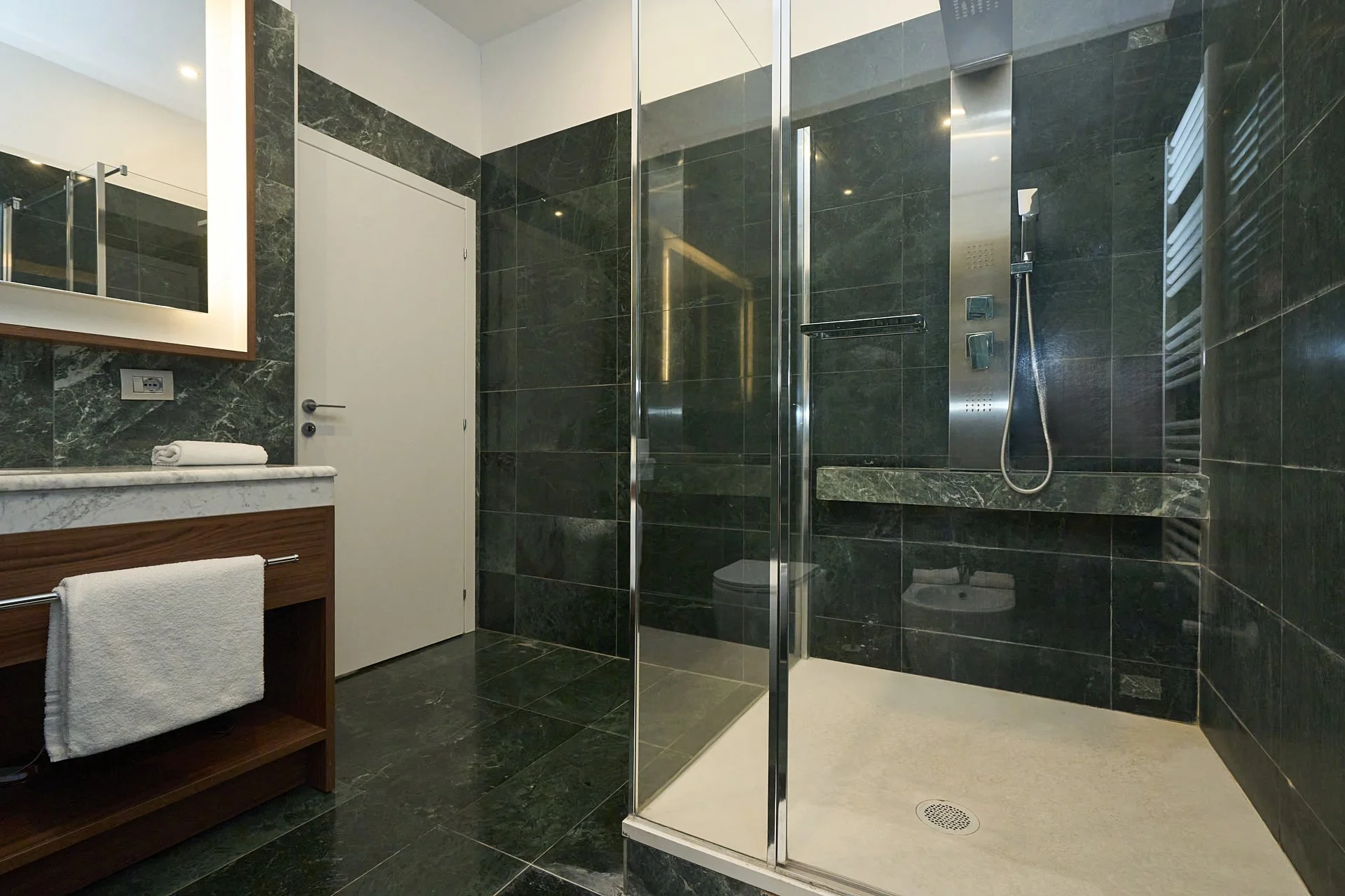 Bagno moderno con pareti e pavimenti in marmo verde scuro, doccia walk-in con vetri trasparenti, lavabo con specchio con luce, asciugamani bianchi.
