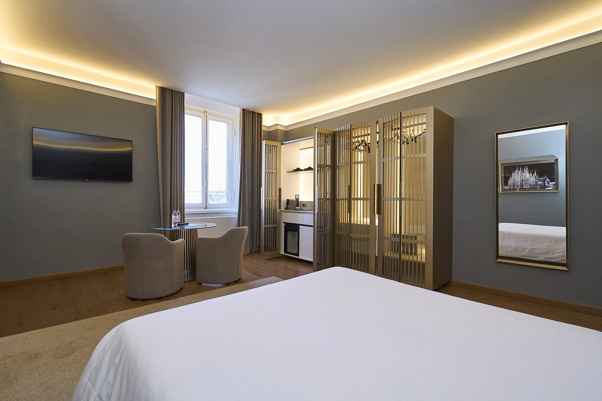 Camera da letto moderna con pareti blu e mobili in legno chiaro. Zona soggiorno con due sedie e un tavolino vicino alla finestra. Rotonda con un televisore a schermo piatto sulla parete. Specchio rettangolare sulla parete opposta.