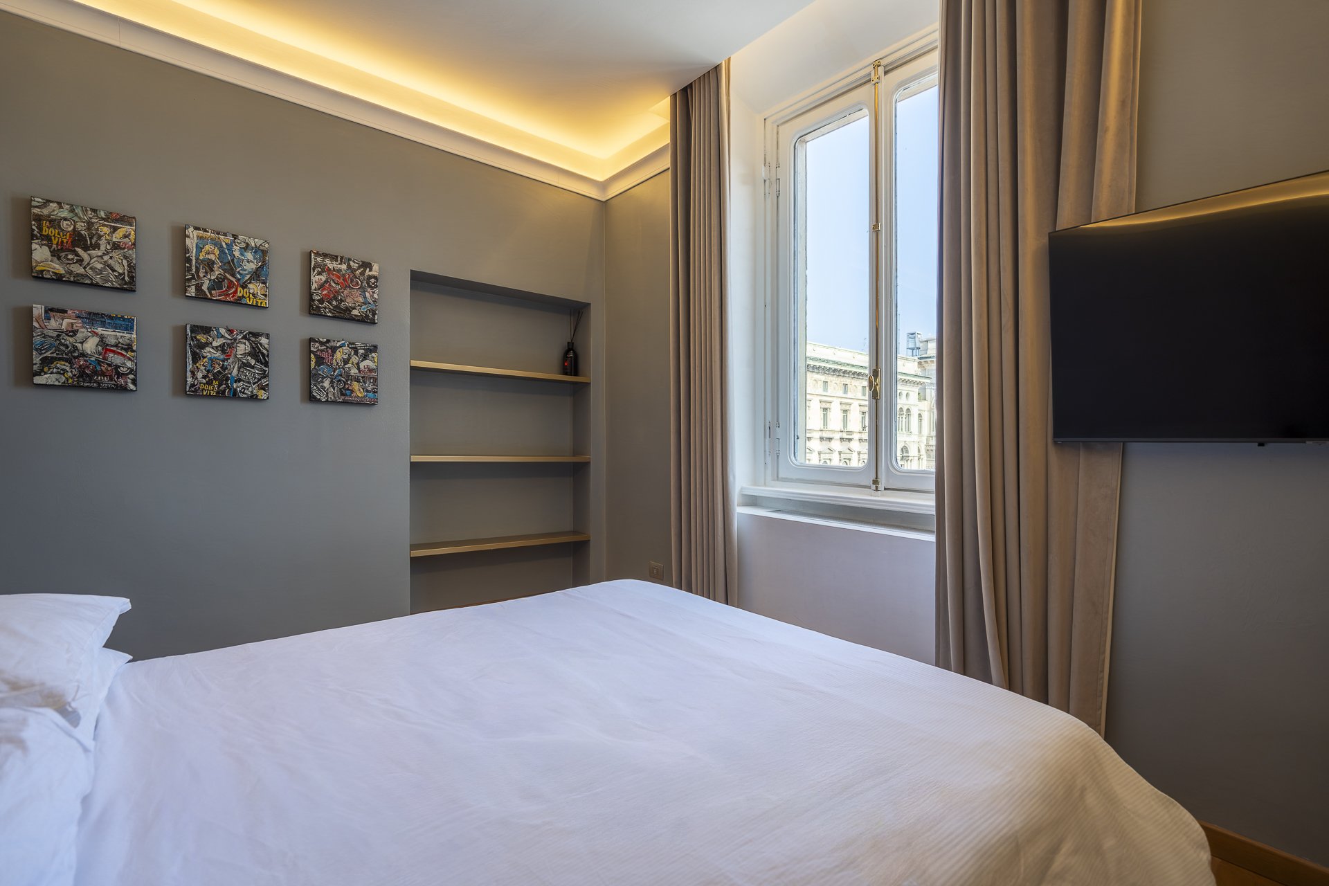 Camera da letto con letto matrimoniale, parete con sei quadri astratti, finestra grande con vista su edifici storici, televisore a schermo piatto, tende beige e illuminazione soffusa a soffitto.