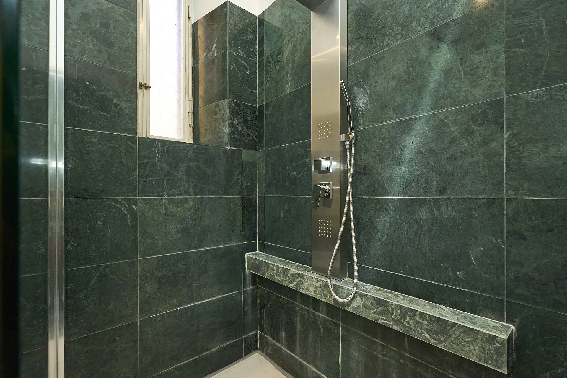 Doccia con pannello in acciaio, maniglia e bocchetta a mano, rivestimento in piastrelle verdi scure, finestra in vetro.