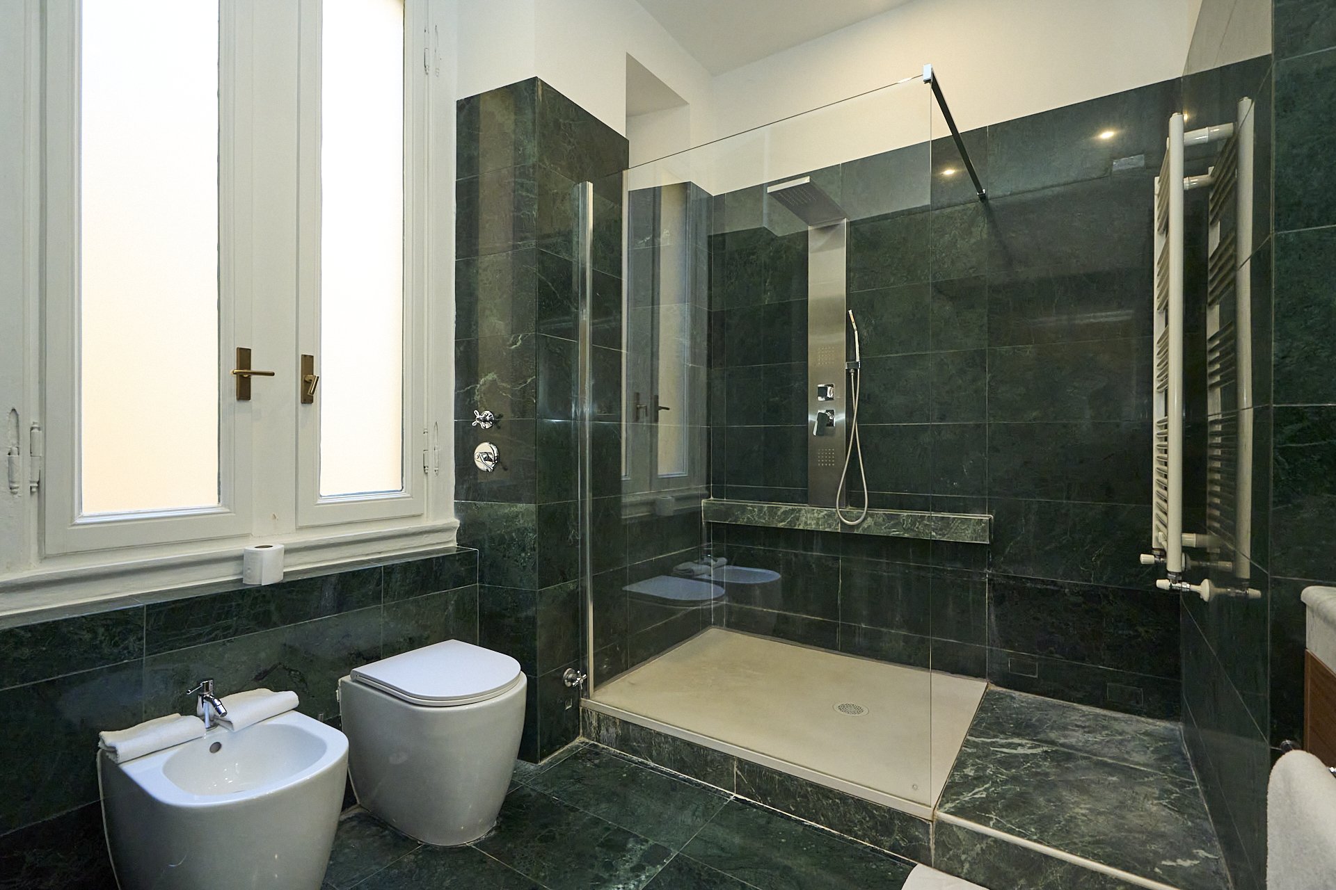 Bagno moderno con pareti e pavimenti in marmo verde scuro, doccia a filo pavimento con vetro divisorio, toilette e bidet bianchi vicino alla finestra con persiane.