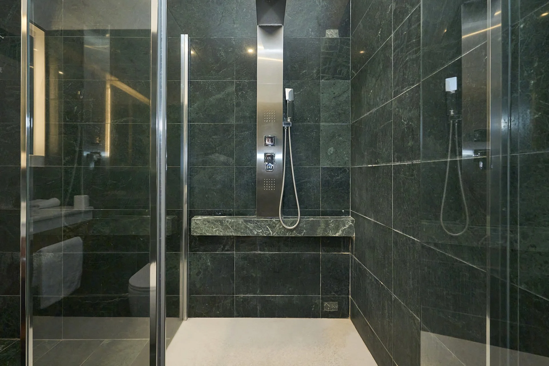 Doccia moderna con pareti in marmo verde scuro, sezionatore in vetro e portascarpe, con soffione e lavandino integrati.